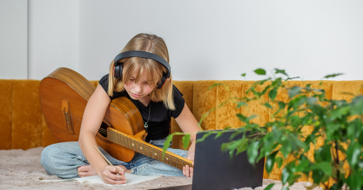 Bambina con cuffie e chitarra mentre prende appunti davanti al laptop, esempio di apprendimento musicale a distanza.